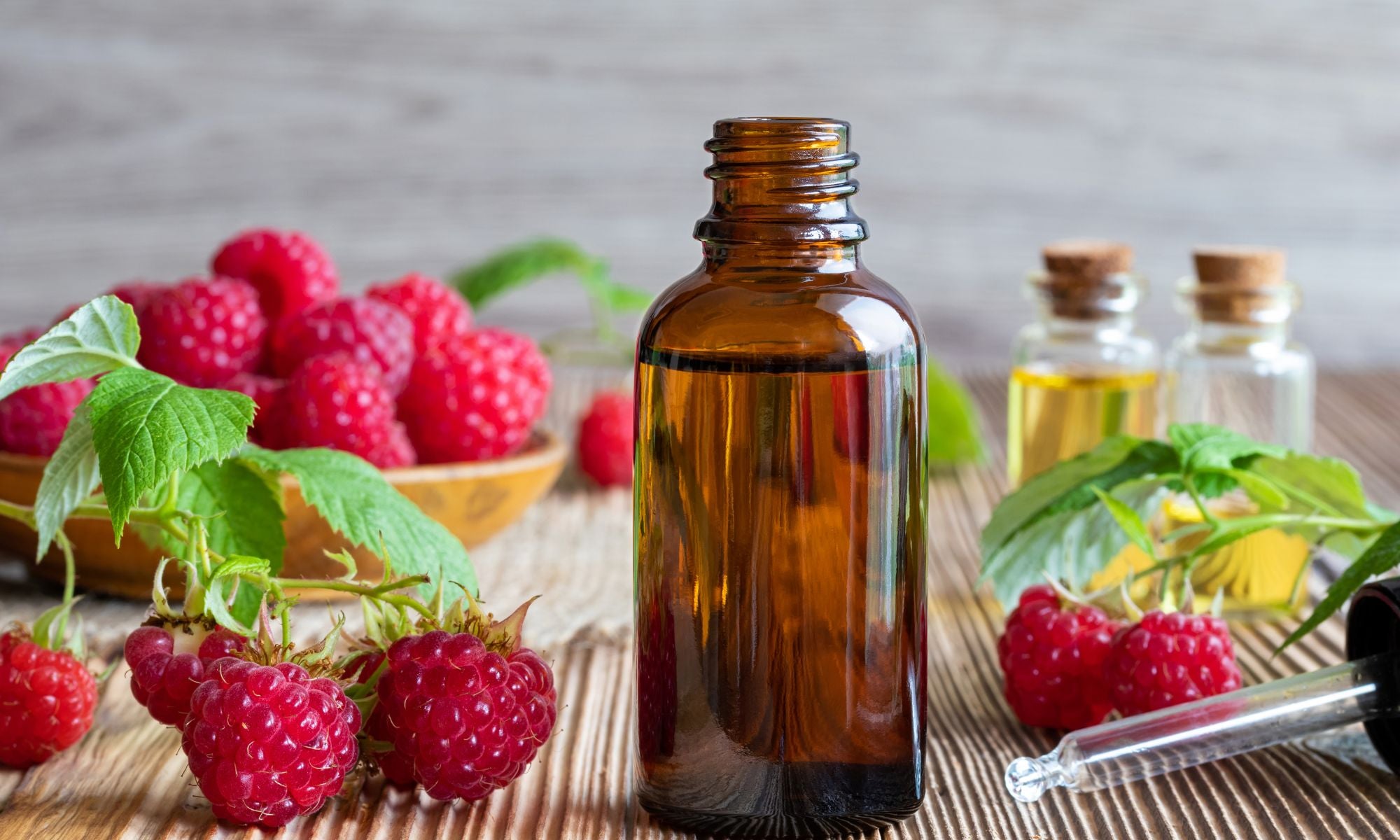 The image presents a still life composition centered around raspberries and what appears to be raspberry seed oil. A large, amber-colored glass bottle dominates the foreground, filled with a dark, golden liquid – likely the oil itself.